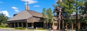 A church with a wooden exterior, sloped roof, and a cross on top stands beside a freestanding wooden bell tower surrounded by trees under a blue sky with scattered clouds.