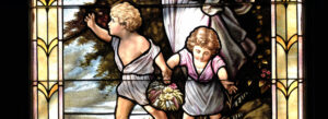 Stained glass window showing two young children holding hands. One child carries a basket of flowers, while the other reaches outward. Trees and a scenic landscape appear in the background.