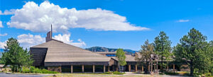 Modern building with a sloped roof and prominent triangular entrance, surrounded by pine trees and mountains under a blue sky with fluffy clouds.