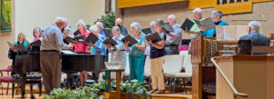 A choir of older adults stands in front of a piano, singing from sheet music while a conductor leads them. An organist plays at the right, and the scene is set in a well-lit church with greenery in the foreground.