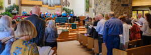 A congregation stands and sings from hymnals in a church sanctuary with wooden pews, a choir in robes at the front, and a stone wall beside the altar decorated with musical instruments.