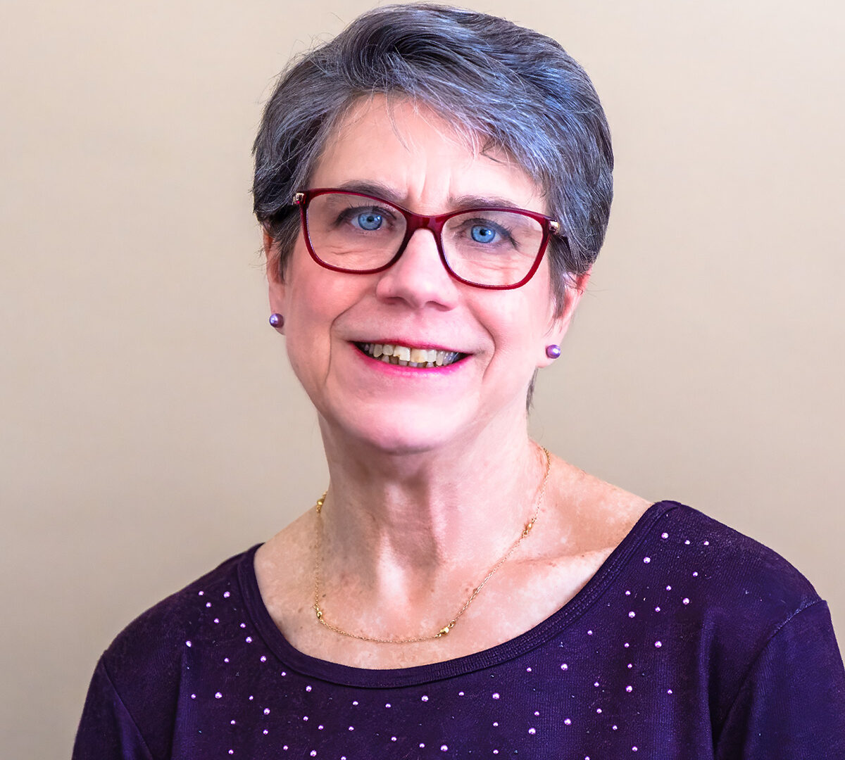 Smiling older woman with short gray hair and red glasses, wearing a dark purple top with small sparkles and a gold necklace, standing in front of a plain beige background.