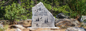 A stone marker with engraved text reading "Kent Dannen Meditation Trail, Presbyterian Community Church of the Rockies," featuring mountain, trees, a deer, and a bird, set among rocks and greenery.